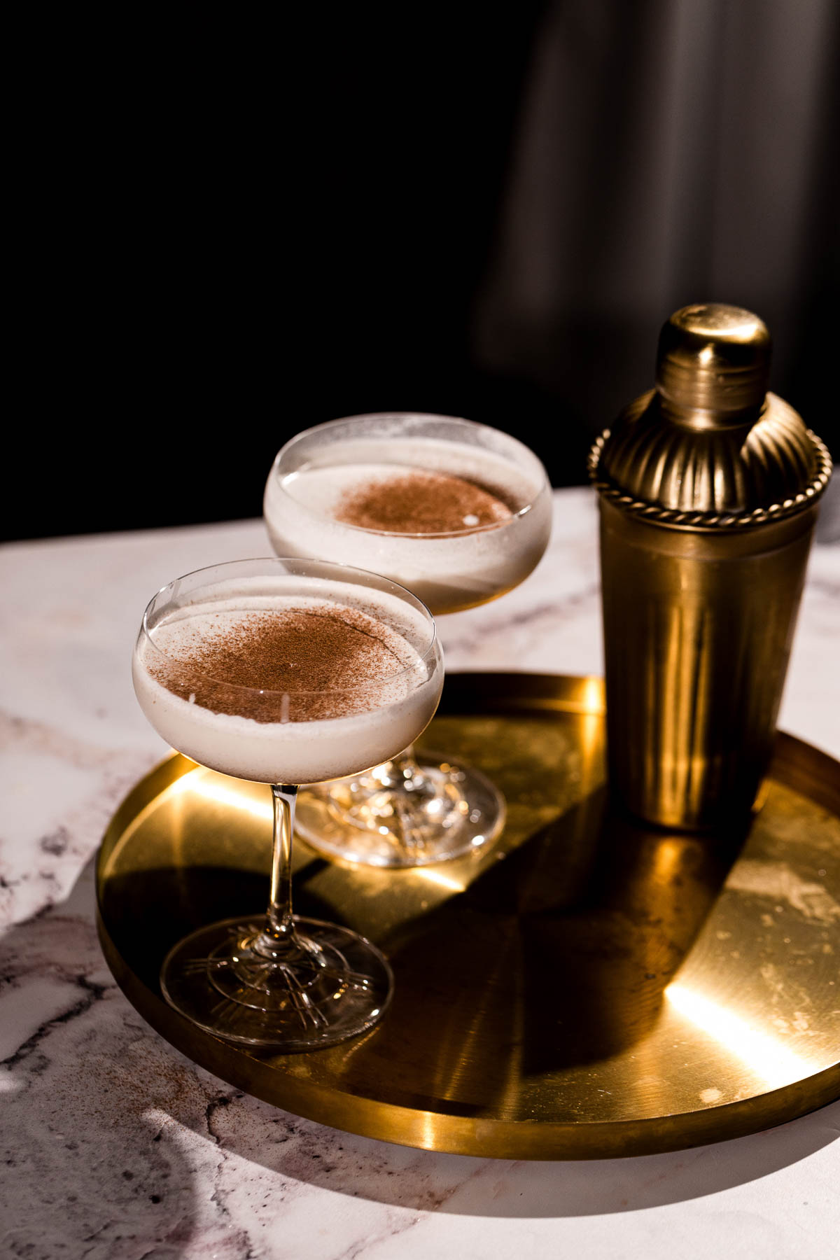 Two cinnamon-topped frothy eggnog martini cocktails sit on a gold tray beside a cocktail shaker on marble.