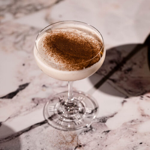 A coupe glass containing a creamy eggnog martini topped with grated nutmeg and cinnamon rests on marble.