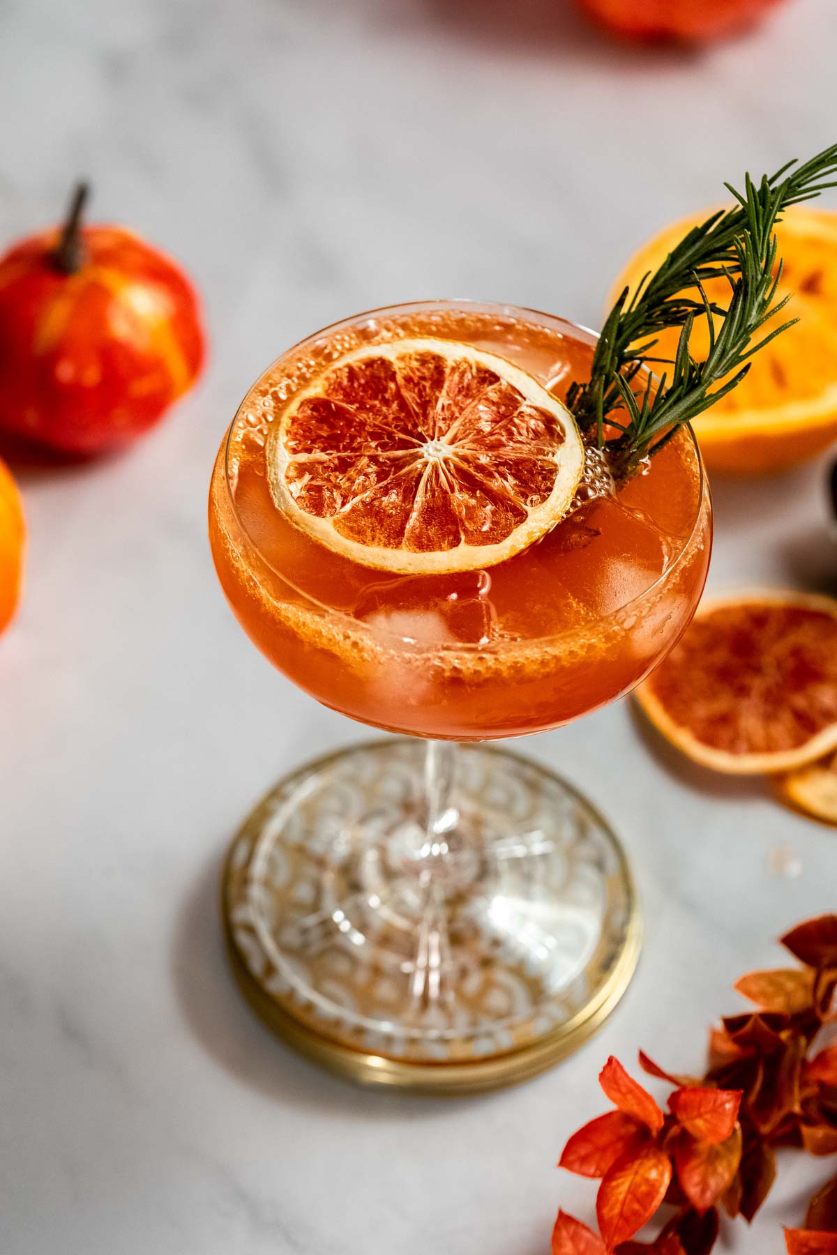 A fall aperol spritz cocktail garnished with dried orange and rosemary sits amidst small pumpkins, dried orange slices, and autumn leaves on marble.