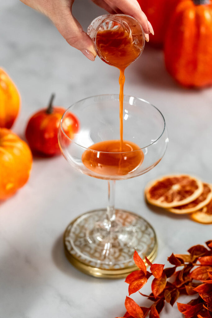 A hand pours pumpkin syrup from a small glass into a coupe glass, surrounded by pumpkins, dried orange slices, and leaves on a white surface.