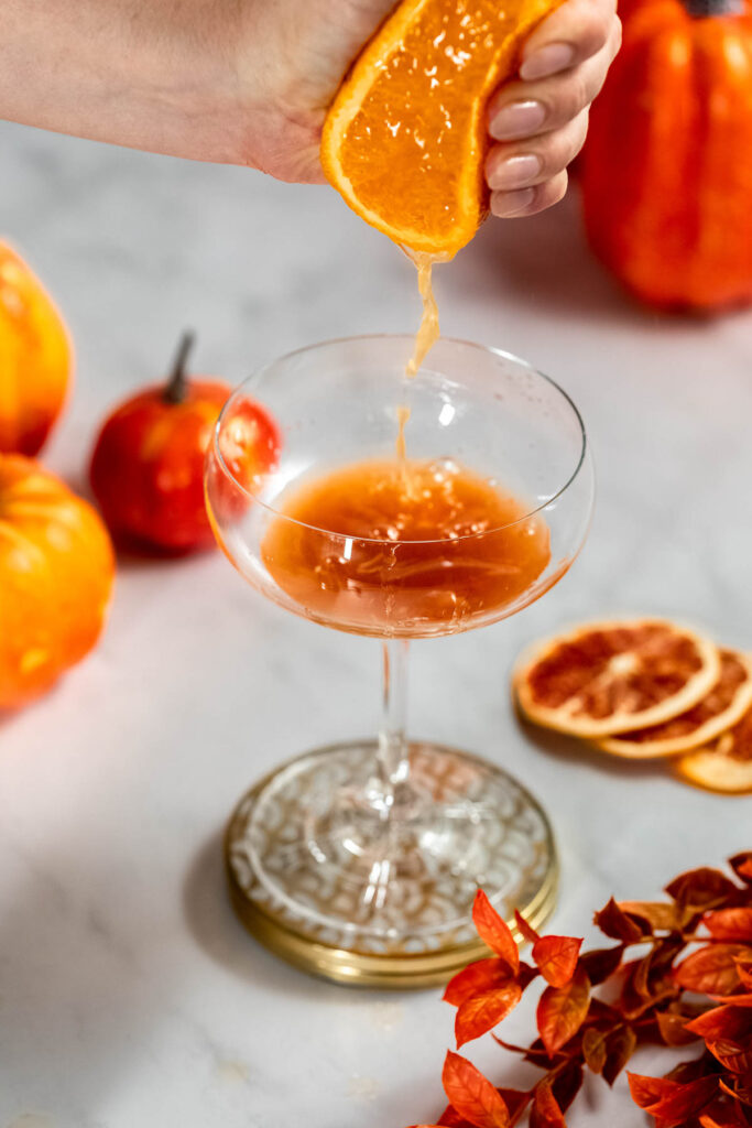 Autumn-themed decor with pumpkins, orange slices, and leaves surrounds a hand squeezing orange juice into a glass.