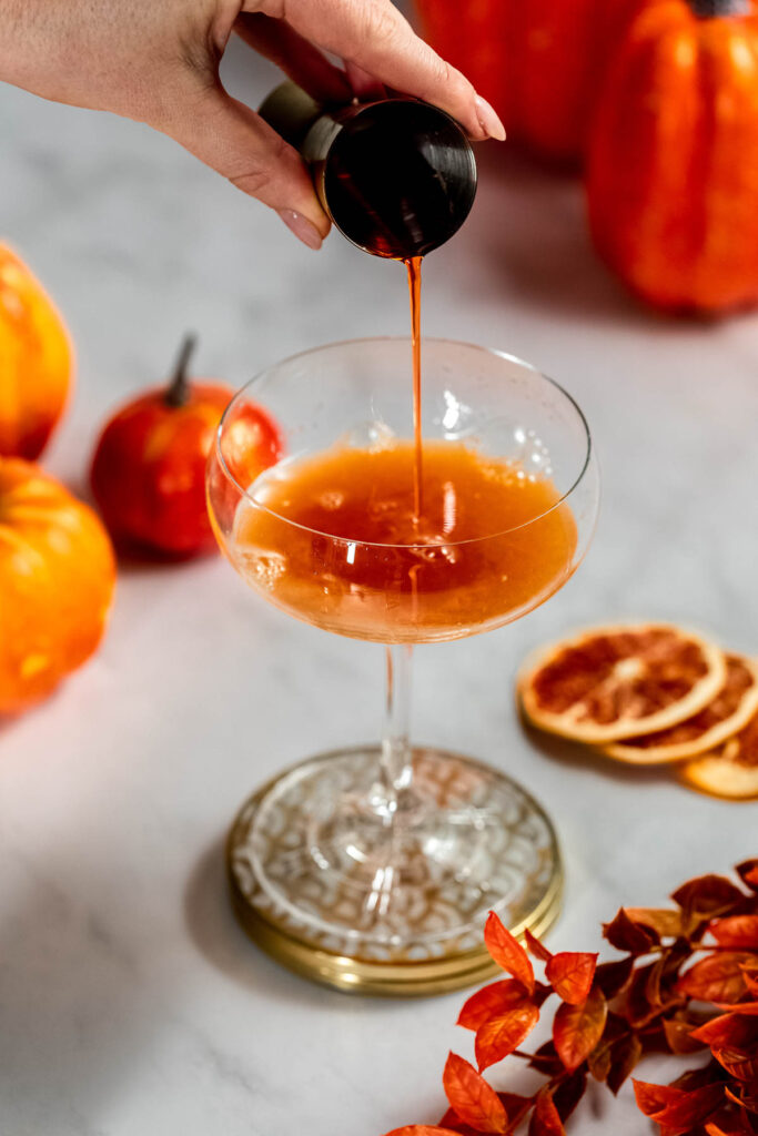 Pouring aperol into a cocktail glass with pumpkins, dried orange slices, and autumn leaves in the background.