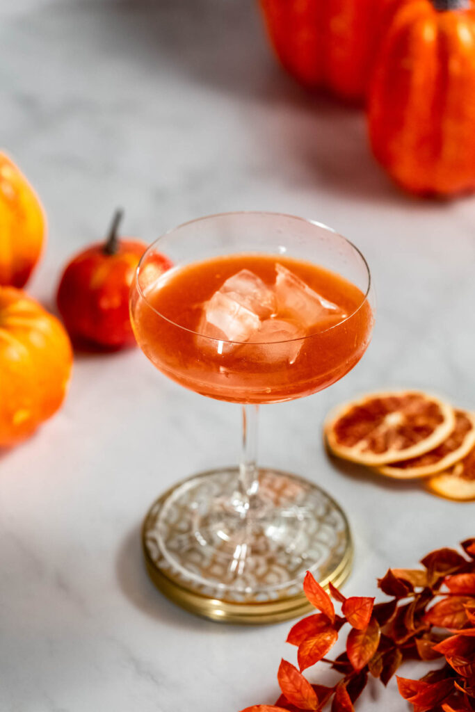 A glass of iced cocktail sits on a marble surface surrounded by small pumpkins and dried orange slices.