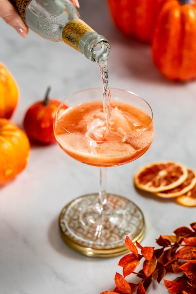 Tonic is being poured into an ice-filled glass, surrounded by pumpkins, dried orange slices, and autumn leaves on a marble surface.