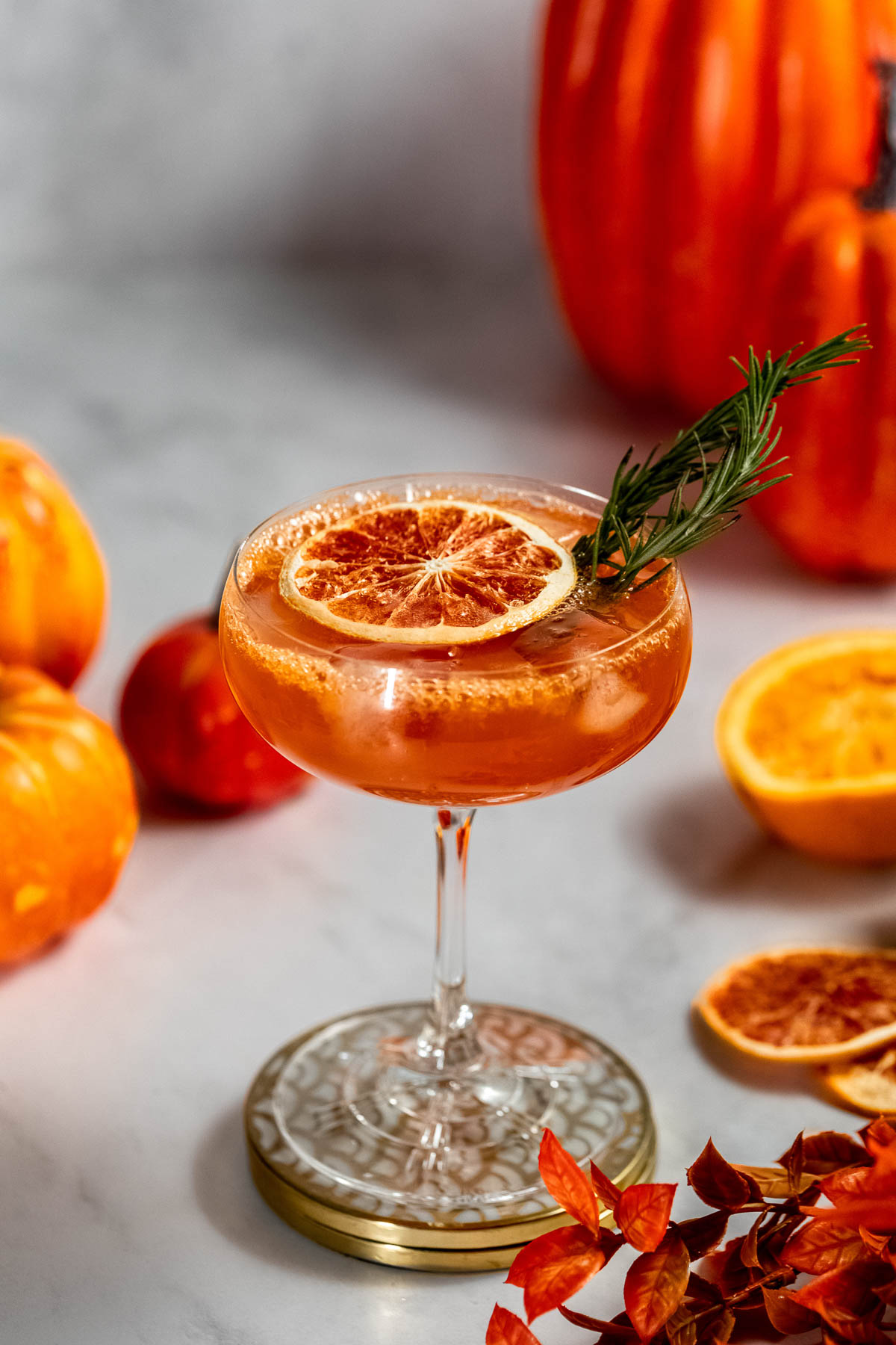 A fall aperol spritz in a wine glass, garnished with a dried orange slice and rosemary sprig, is surrounded by pumpkins, orange halves, and autumn leaves on a marble surface.