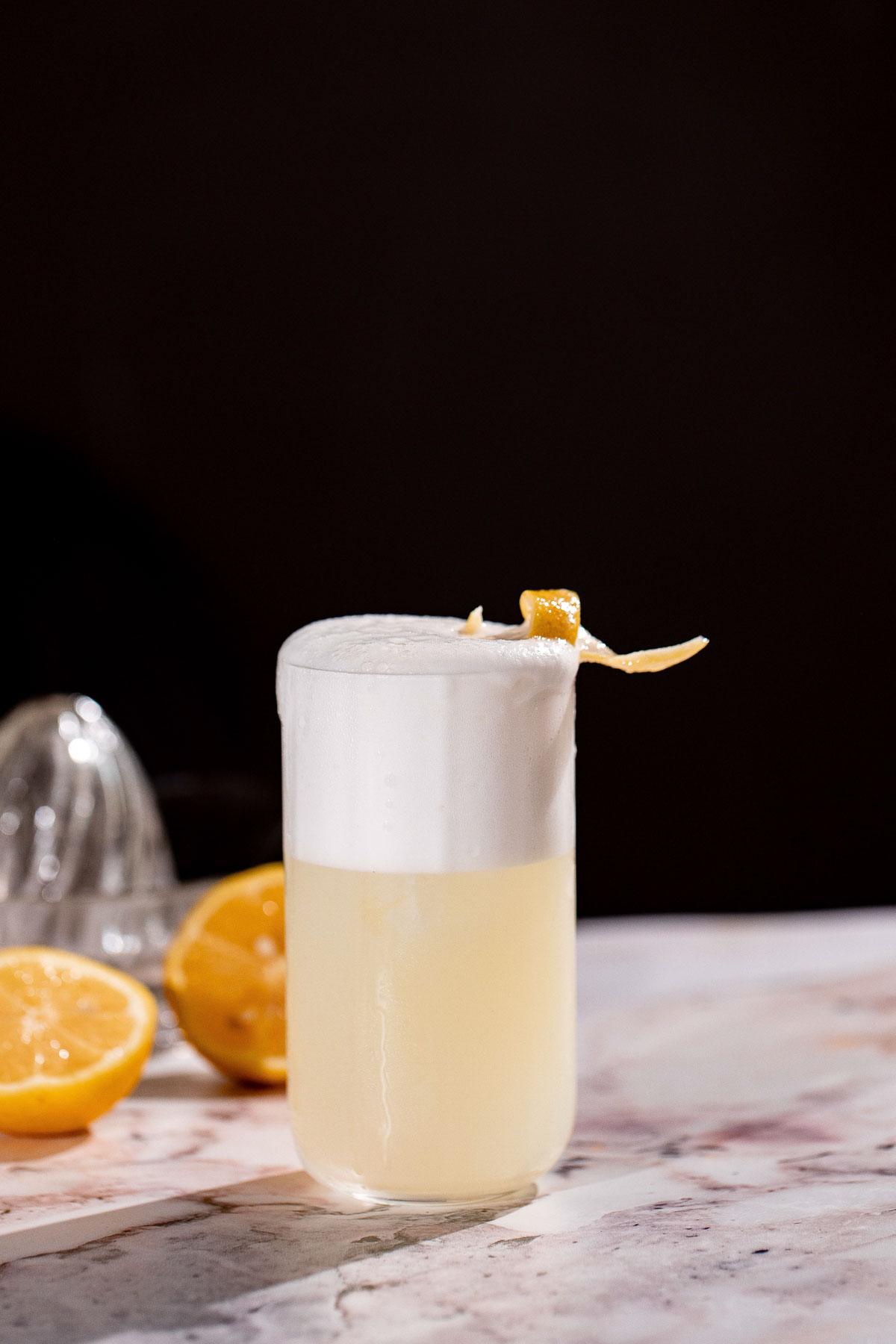 A frothy yellow gin fizz cocktail with a lemon twist sits on a marble surface, surrounded by halved lemons and a juicer.