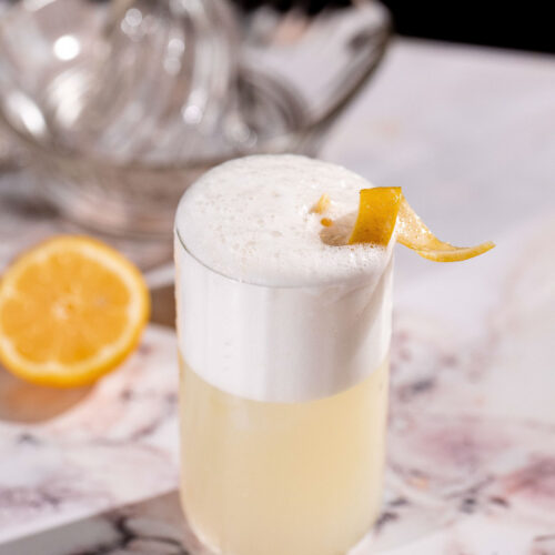 A yellow gin fizz cocktail with a foamy top and lemon peel garnish sits on a marble surface, accompanied by half a lemon and a glass bowl in the background.