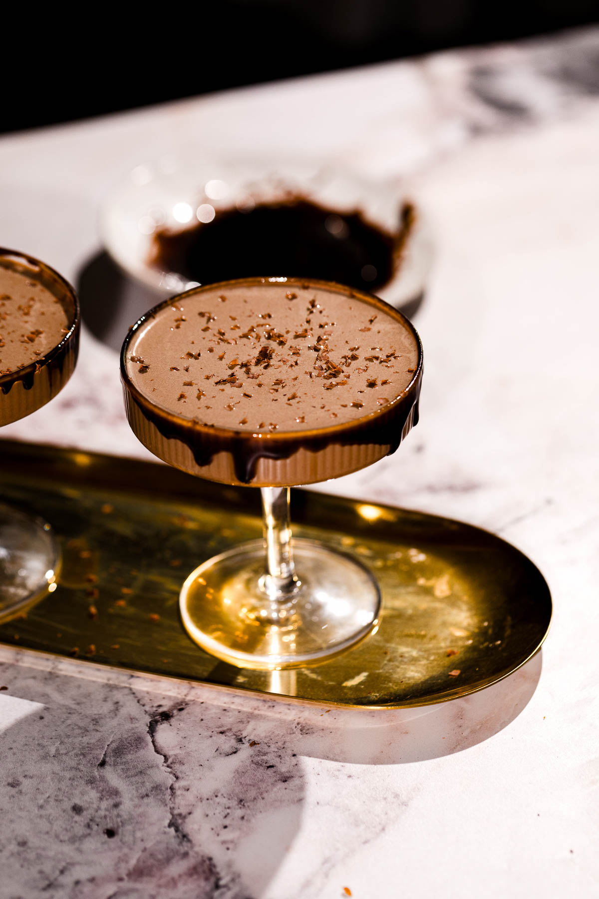 A chocolate martini with chocolate shavings in a coupe glass sits on a gold tray atop a marble surface.