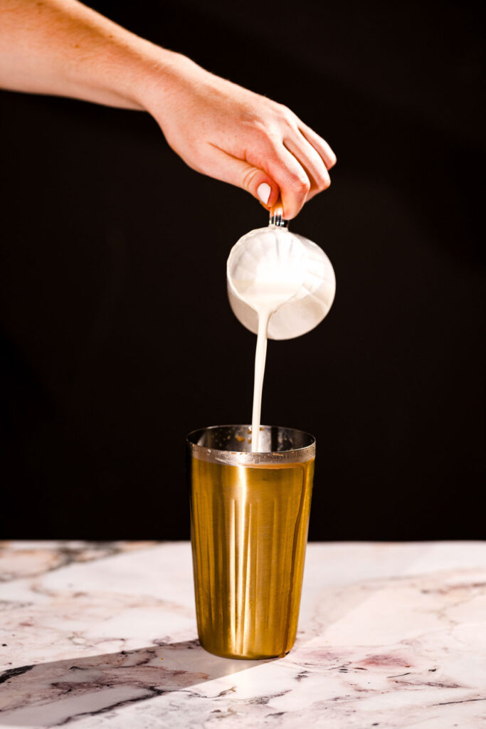 Milk is poured from a white pitcher into a tall gold metal cocktail shaker on a marble surface.