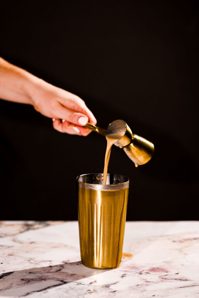 A hand pours chocolate liquor from a measuring cup into a metallic shaker on marble.