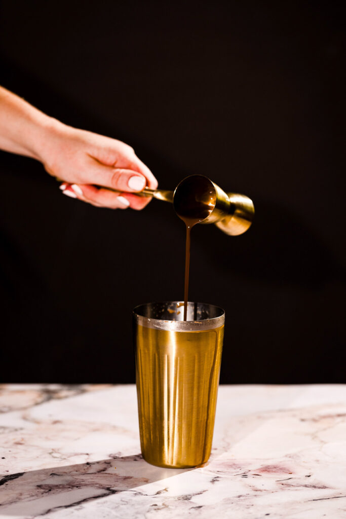 Cream de cocoa is poured from a golden measuring cup into a golden shaker on a marble surface.