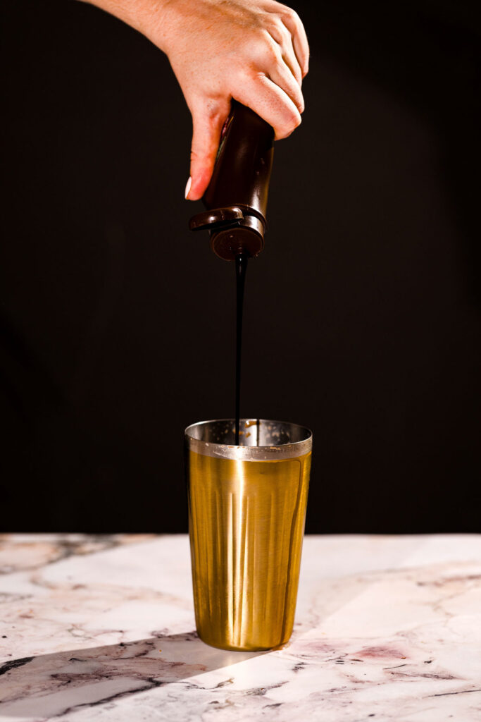 Pouring dark chocolate syrup into a brass cup on a marble surface.