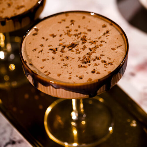 Two glasses of chocolate martinis are topped with chocolate shavings on a tray.