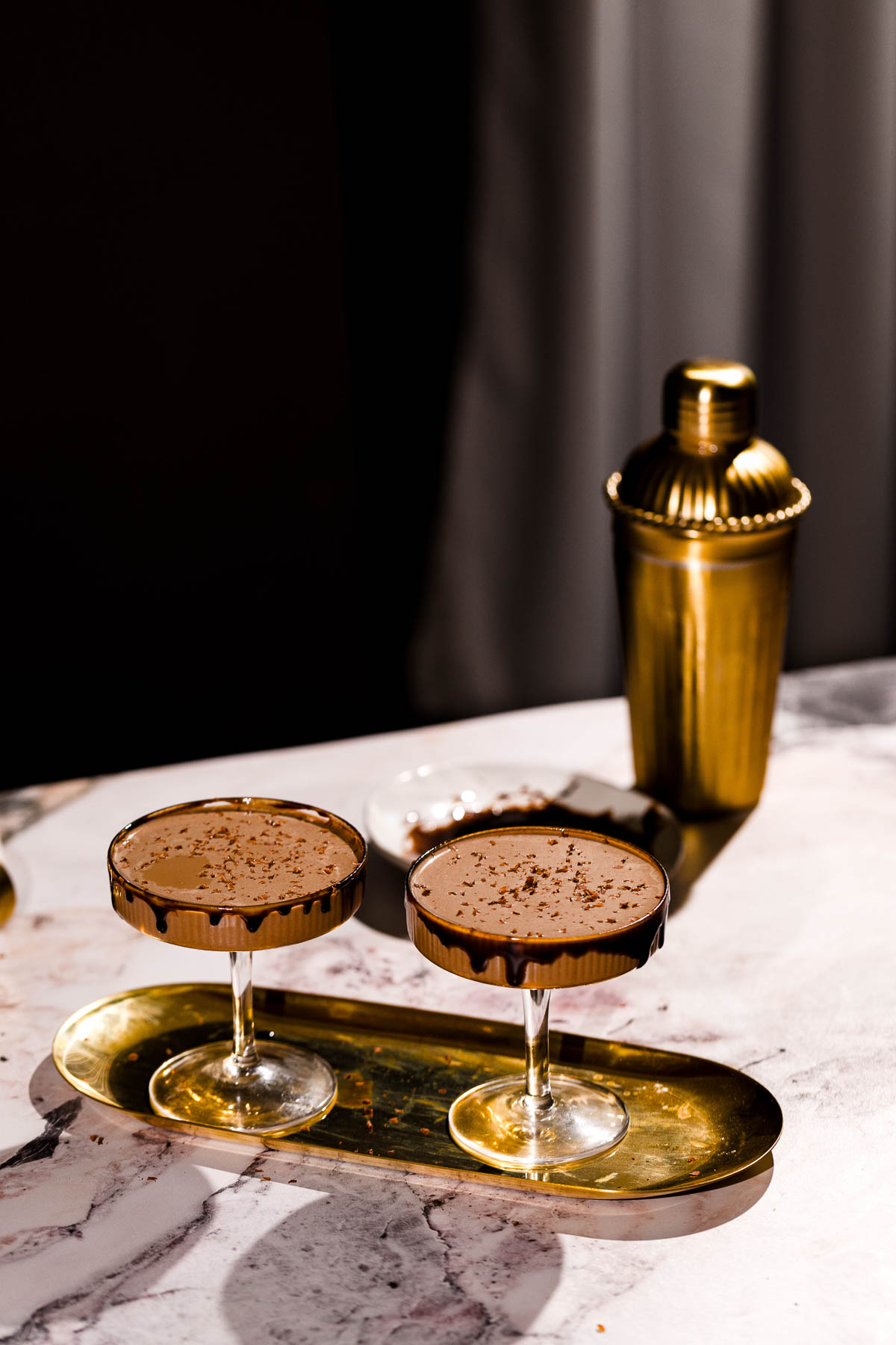 On a marble surface, two chocolate martinis in coupe glasses sit on a gold tray with a gold cocktail shaker in the background.