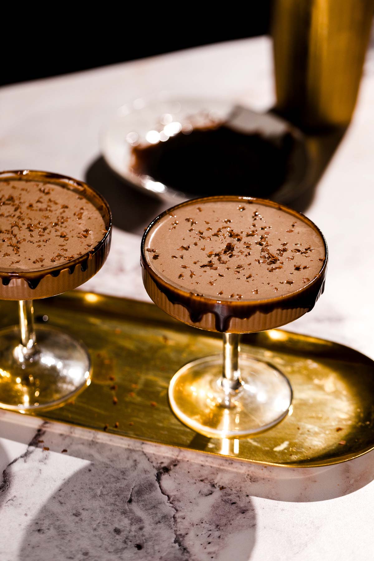 Two chocolate martinis sit on a gold tray with a shaker and chocolate powder in the background.