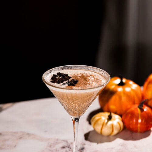 A martini glass with a creamy pumpkin pie martini and chocolate shavings sits on a marble surface with decorative pumpkins in the background.