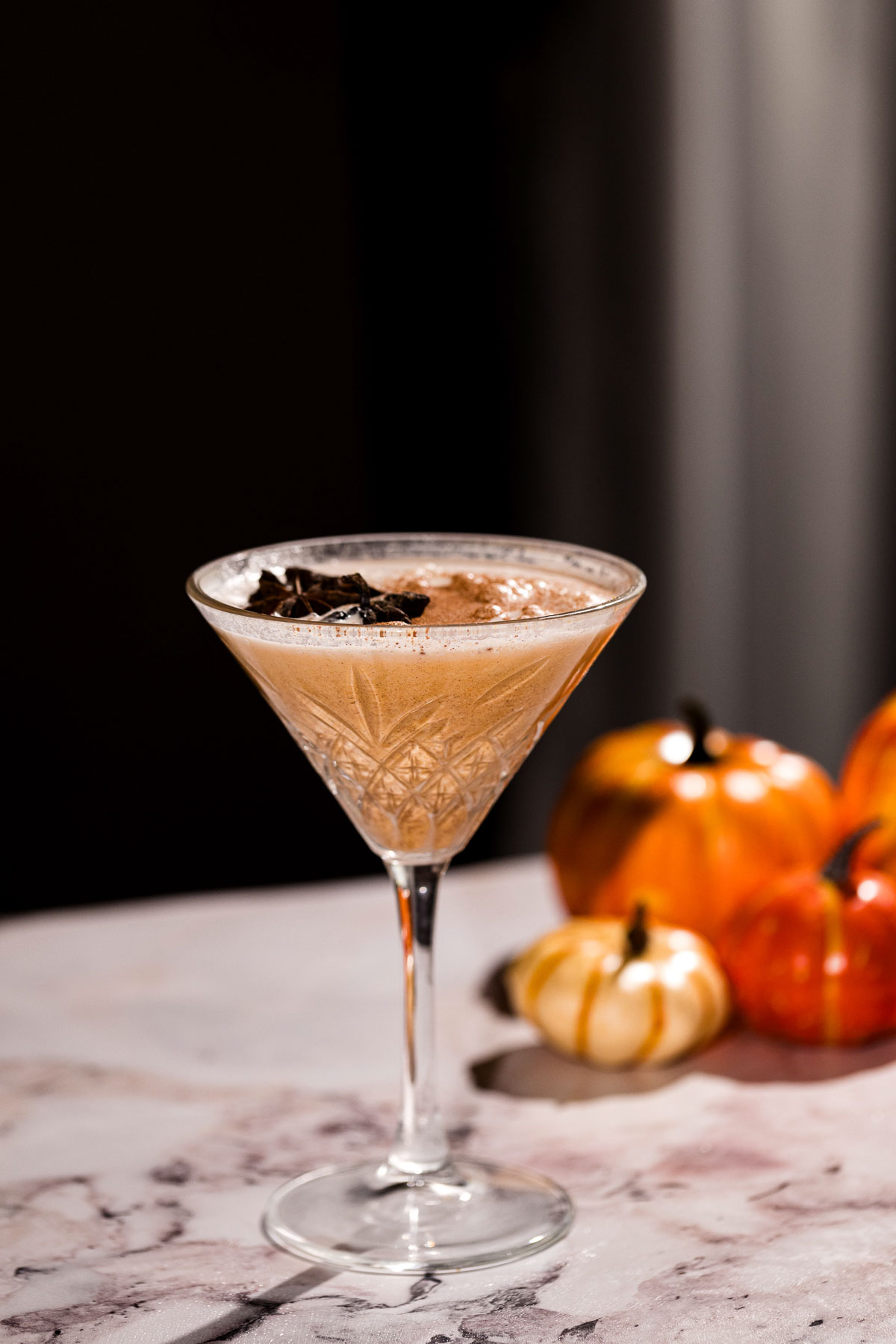 A pumpkin pie martini cocktail in a martini glass, topped with chocolate shavings, sits on a marble surface with small pumpkins in the background.