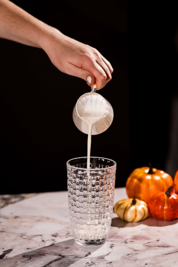 Milk is poured from a jug into a cocktail shaker on a marble surface, with small pumpkins arranged nearby.