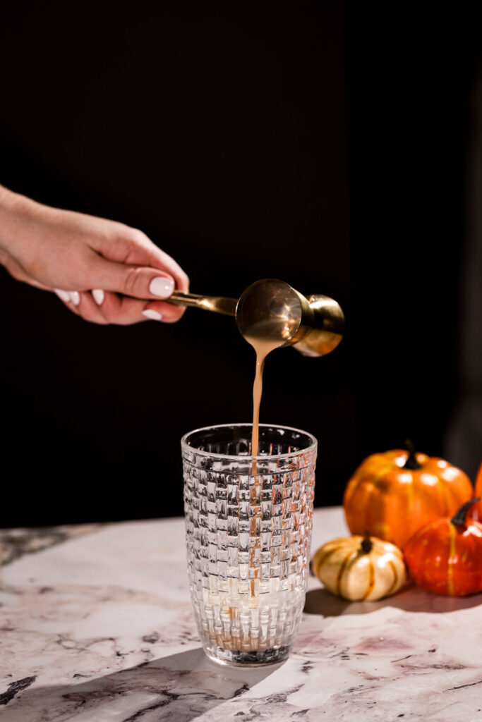 A hand pours irish cream from a brass jigger into a cocktail shaker beside three small pumpkins on the marble countertop.