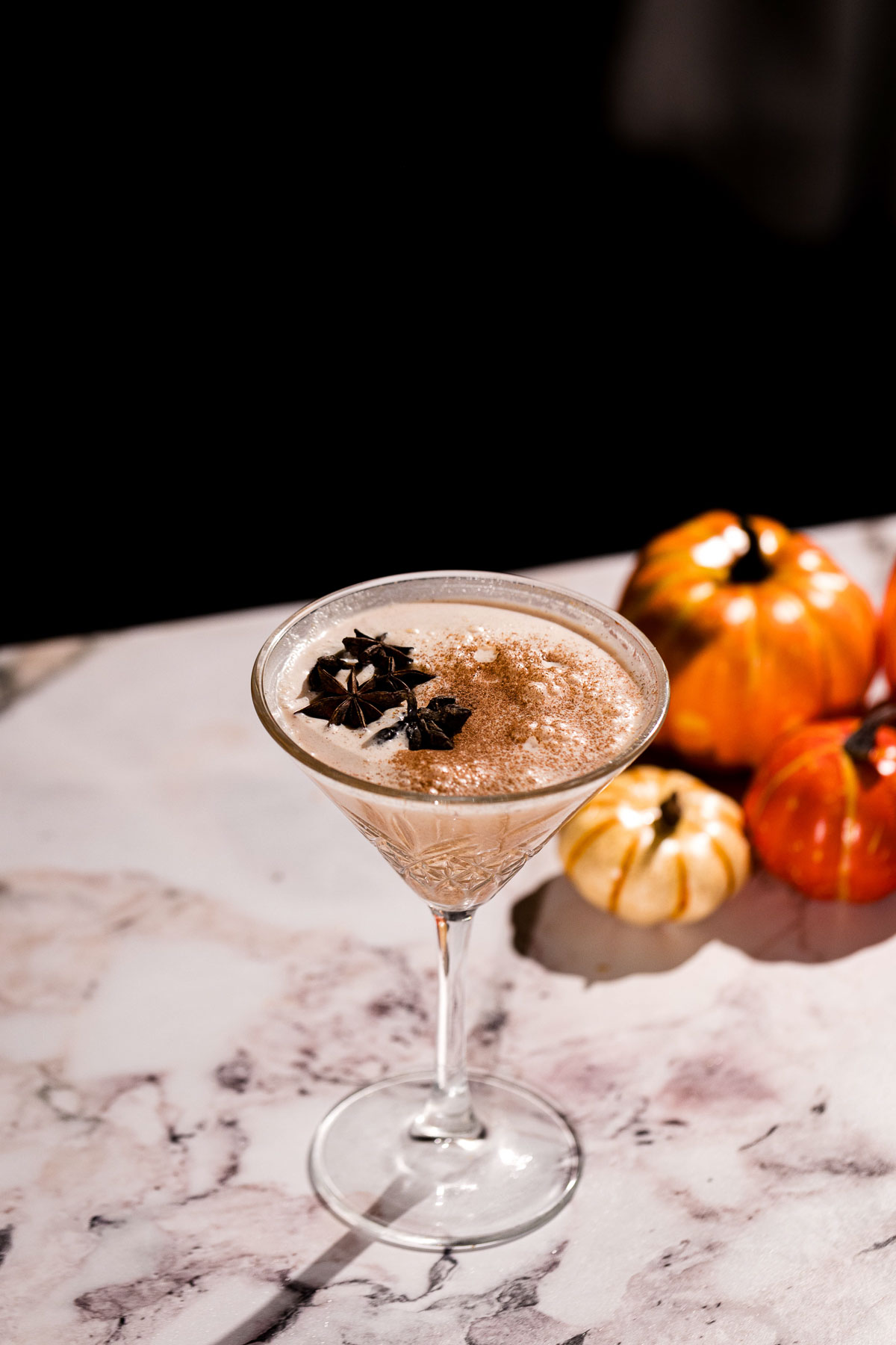 A martini glass filled with a pumpkin pie martini topped with star anise sits on a marble surface beside decorative pumpkins.