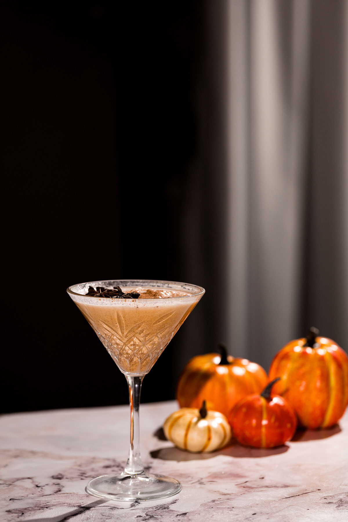 A spiced pumpkin pie martini in a glass sits on a marble surface, adorned with small decorative pumpkins.