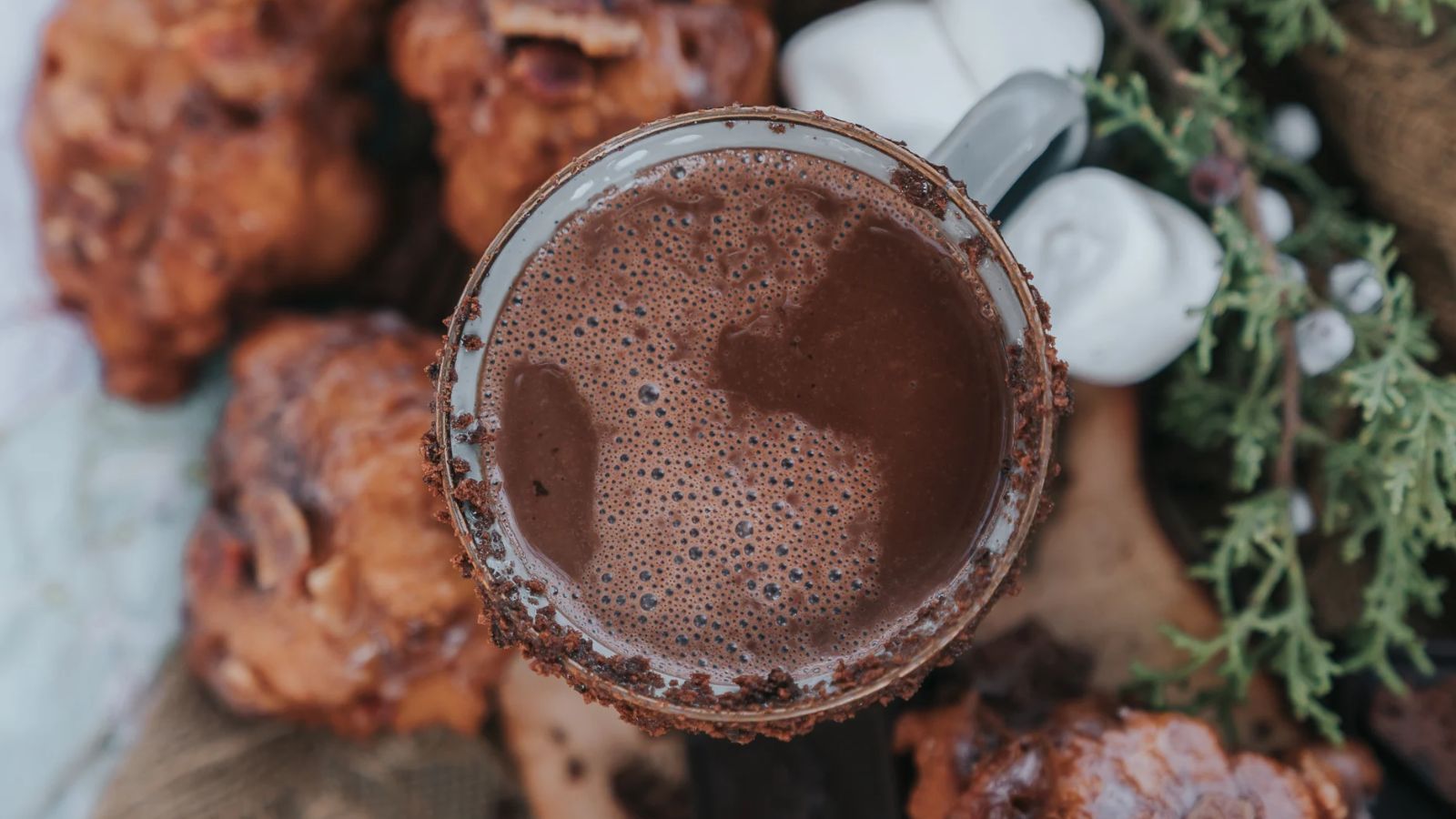 A mug of hot chocolate surrounded by marshmallows and pastries sits against a backdrop of greenery.