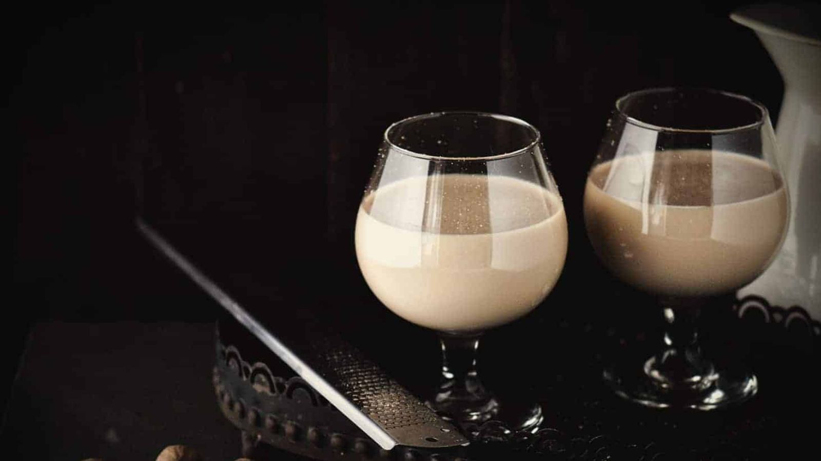 Two brandy snifters contain a creamy beverage on a dark tray beside a grater.