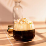 A frothy Cafe Caribbean coffee drink with sprinkles in a clear mug sits on a tiled surface, with a blurred bottle in the background.
