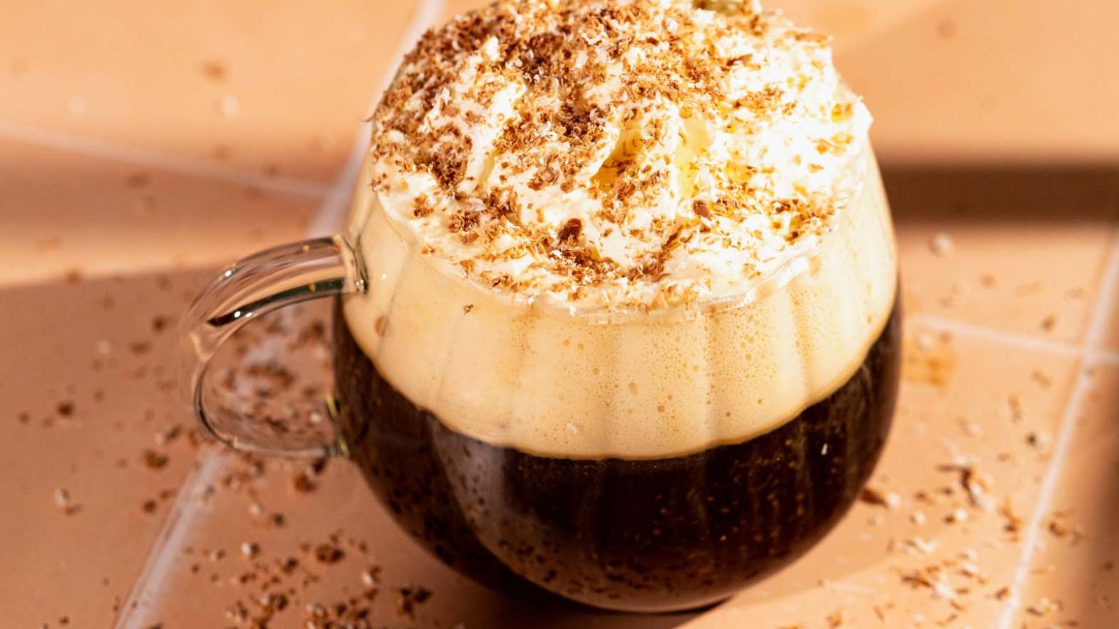 A glass mug of coffee topped with whipped cream and chocolate shavings sits on a tiled surface.