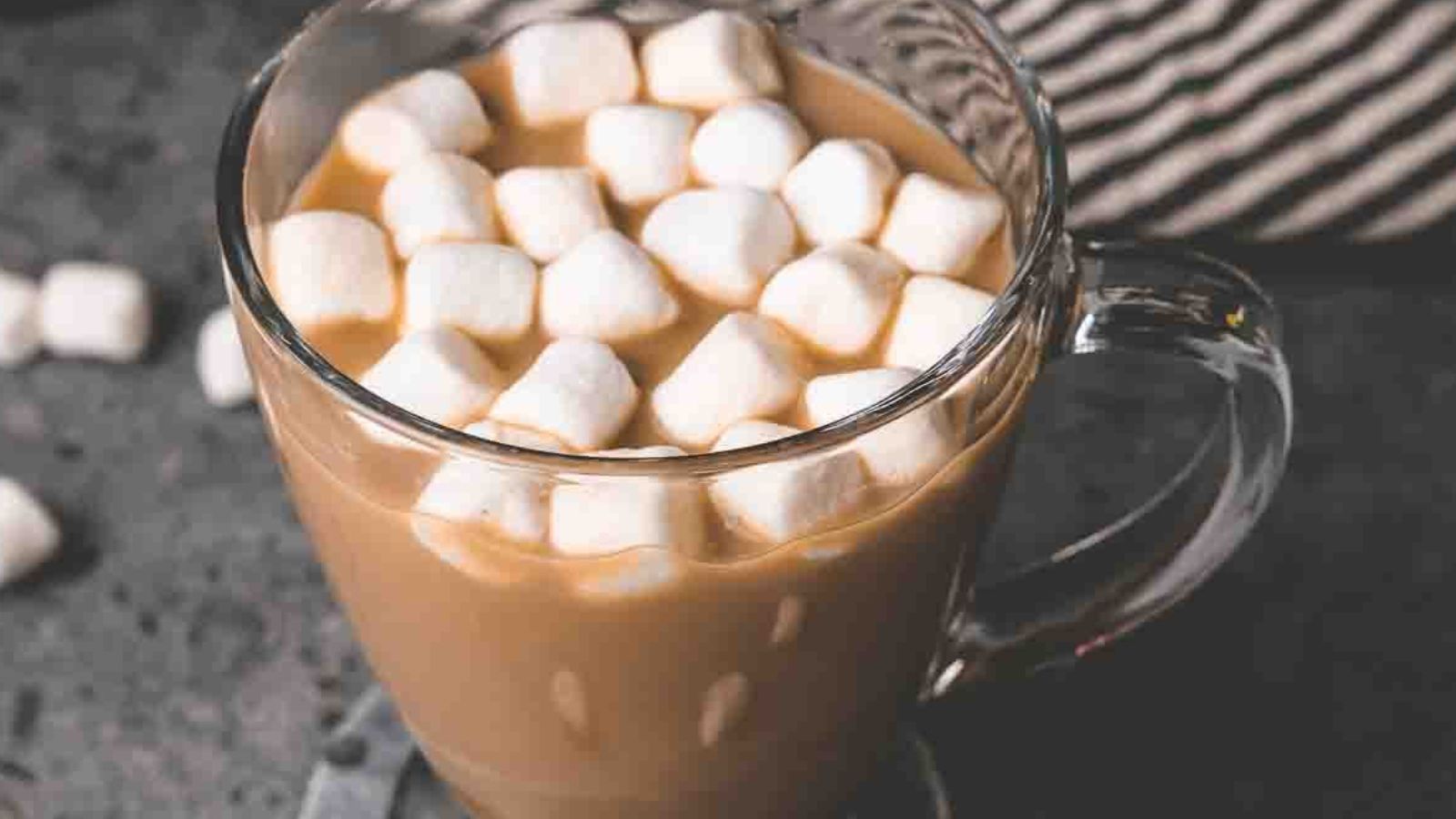 A glass mug of hot chocolate with mini marshmallows sits on a dark surface.