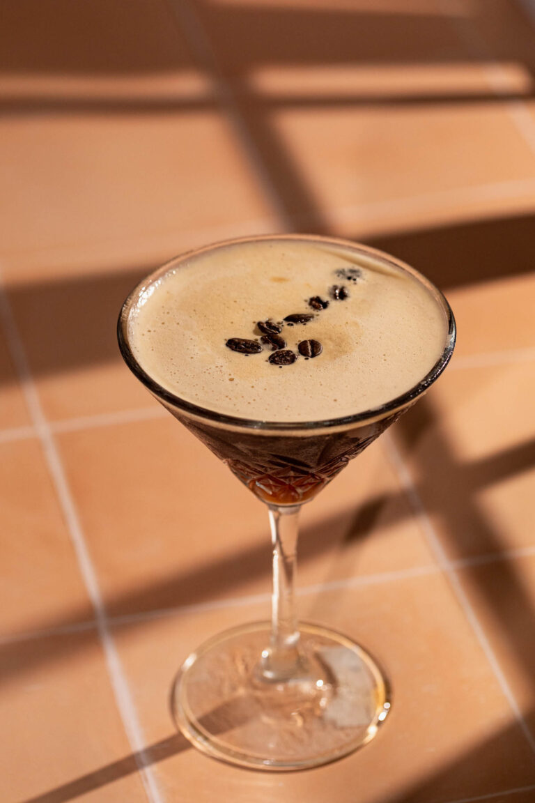 A frothy espresso martini garnished with coffee beans sits on a tiled surface.