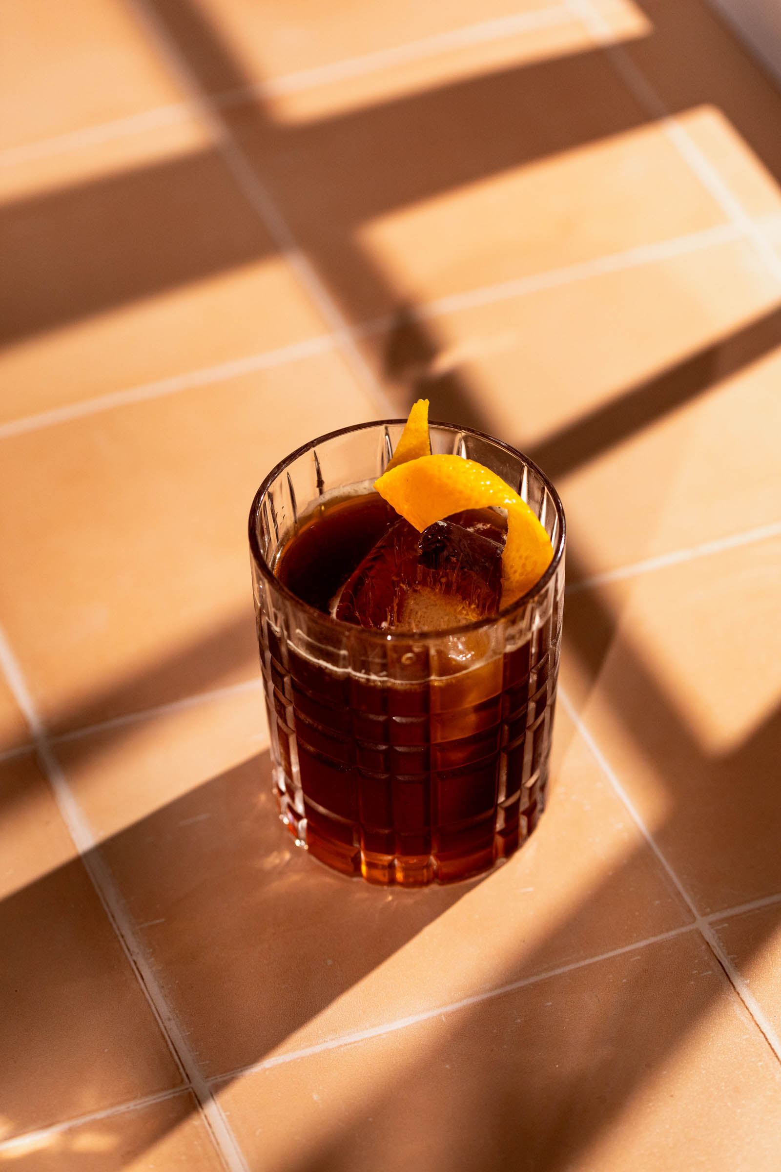 A glass of dark iced espresso old fashioned garnished with an orange peel sits on a tiled surface with shadows.