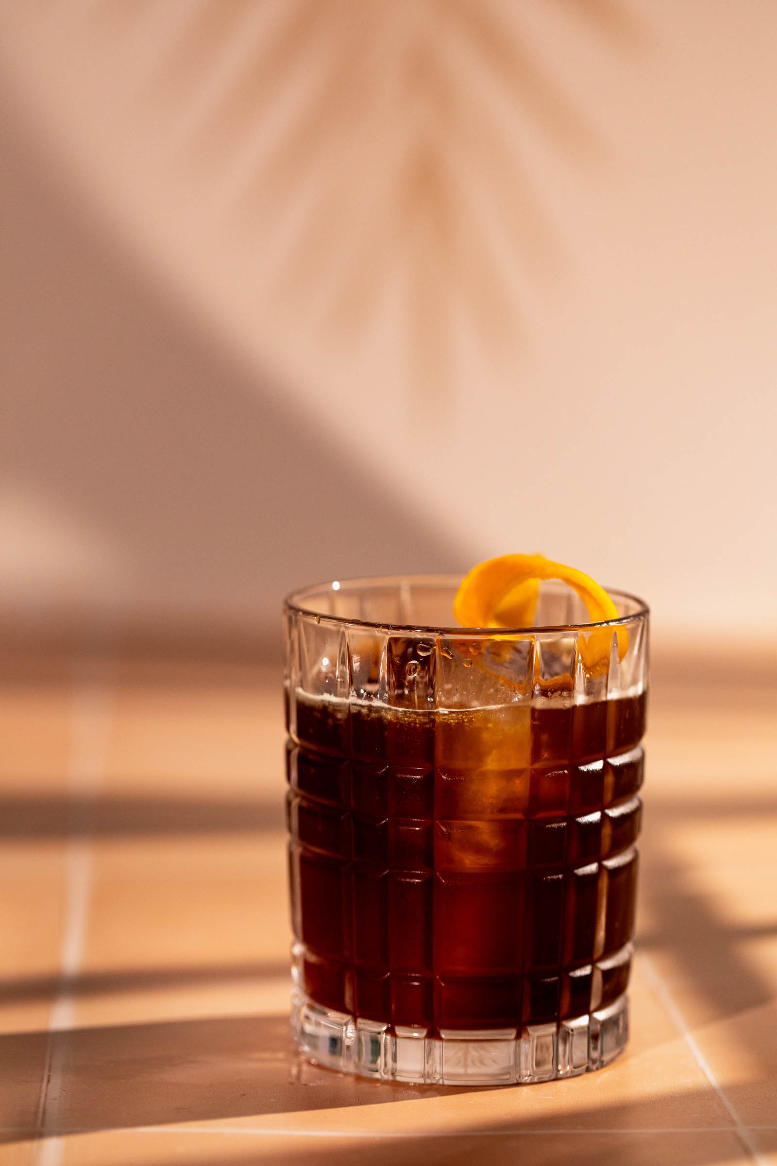 A glass of dark espresso old fashioned with an orange twist sits on a table adorned with soft shadows.