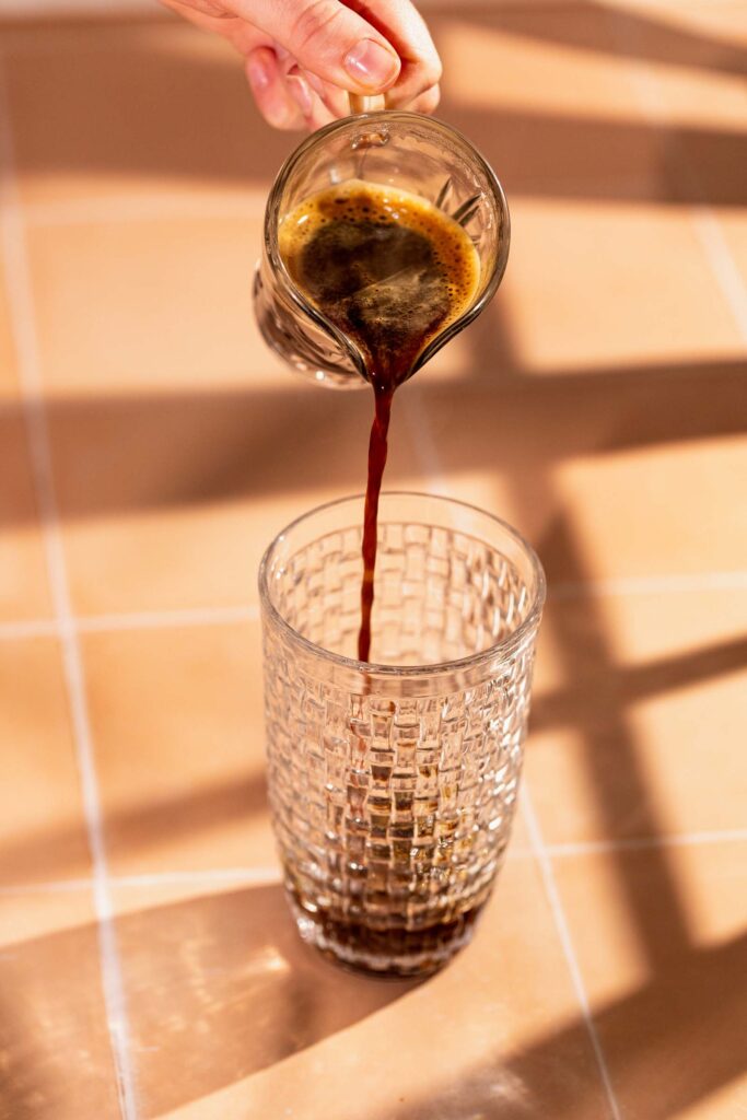 Pouring coffee from a small carafe into a glass on a tiled surface.