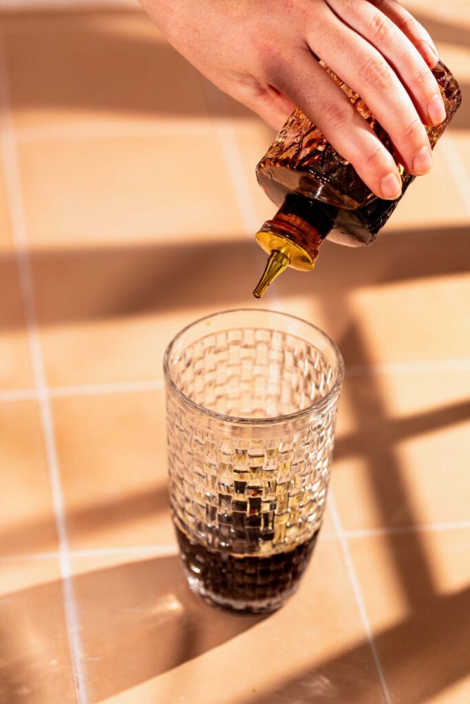 A hand pours bitters from a bottle into a clear patterned glass on a tiled surface.