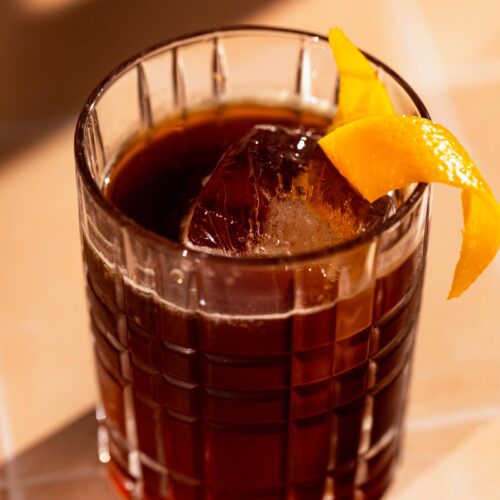 An espresso old fashioned with a large ice cube and orange peel garnish sits on a tiled surface.