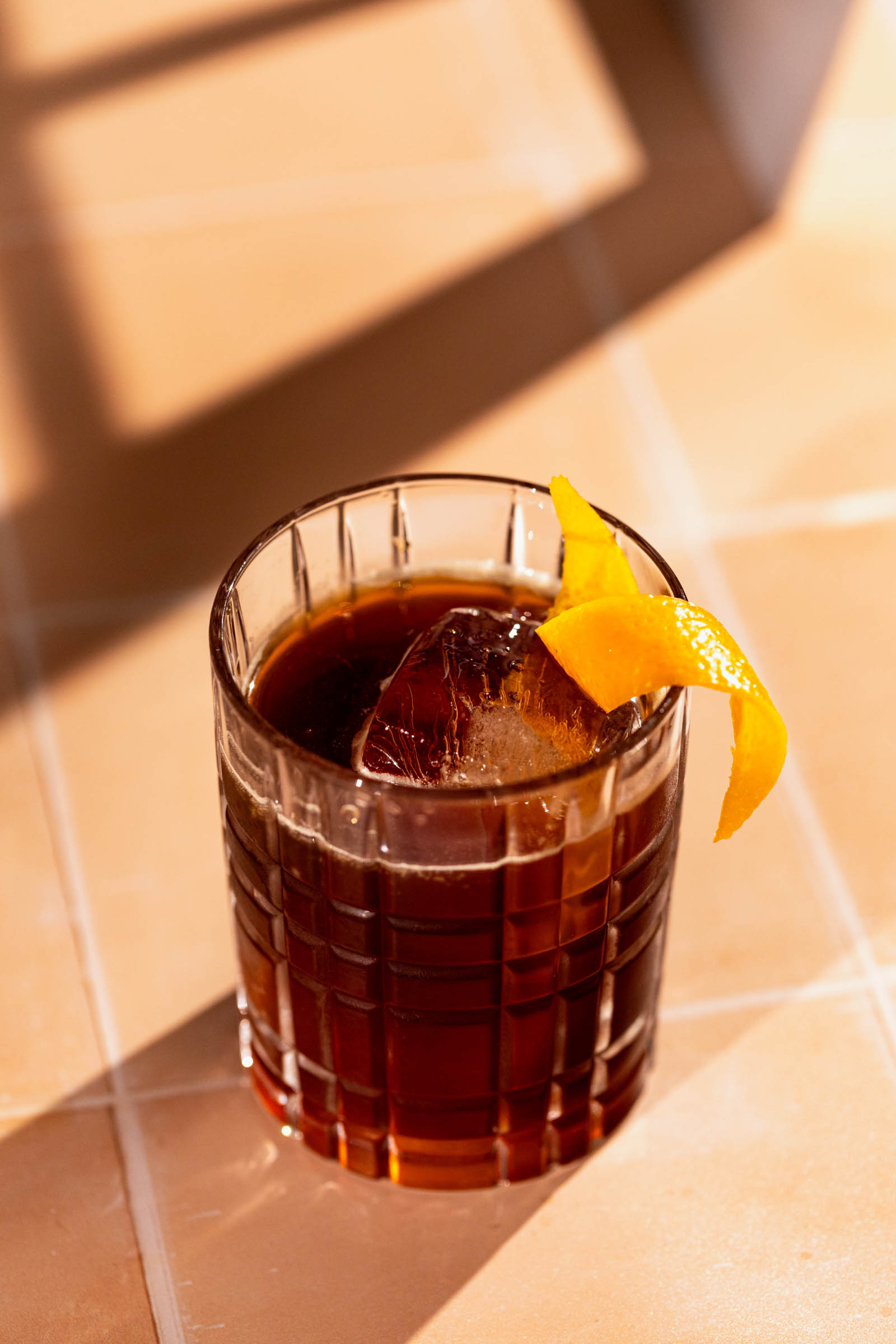 An espresso old fashioned with a large ice cube and orange peel sits on a tiled surface.