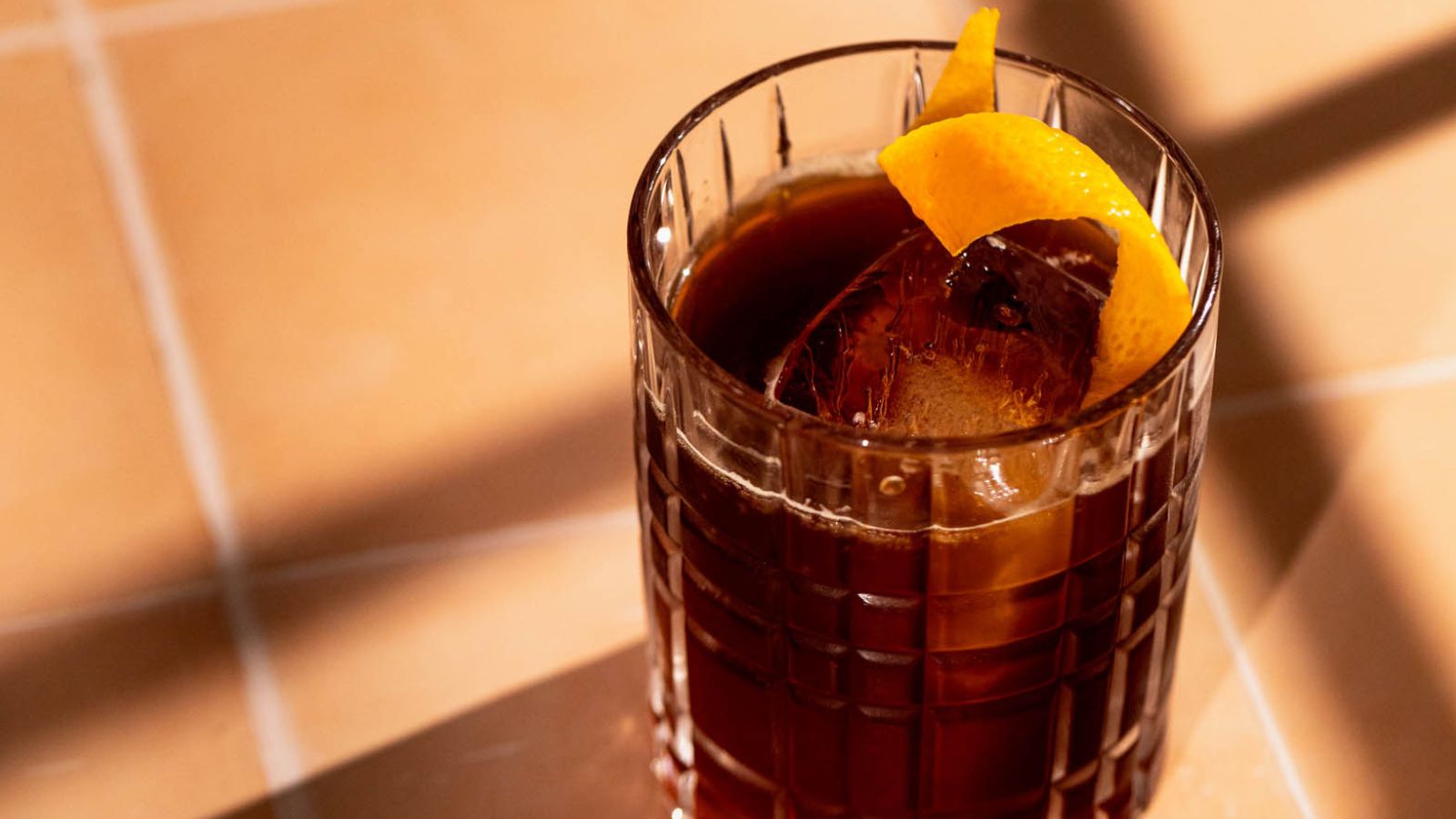 A dark iced drink with an orange twist garnish sits on a tiled surface.