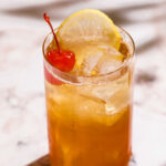 A gin sling in a tall glass with a lemon slice and cherry on top sits on a marble surface.