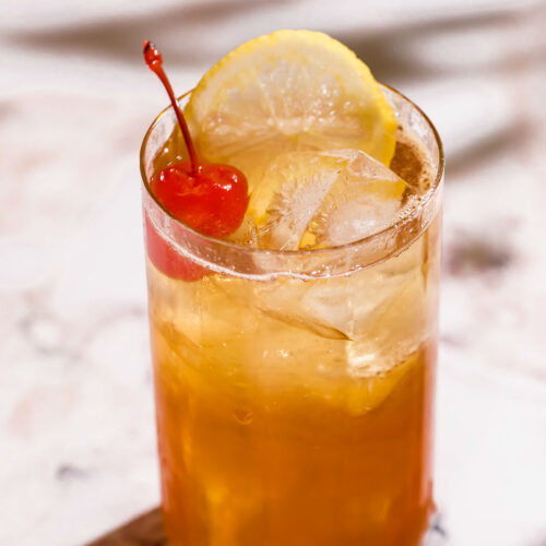 A gin sling in a tall glass with a lemon slice and cherry on top sits on a marble surface.