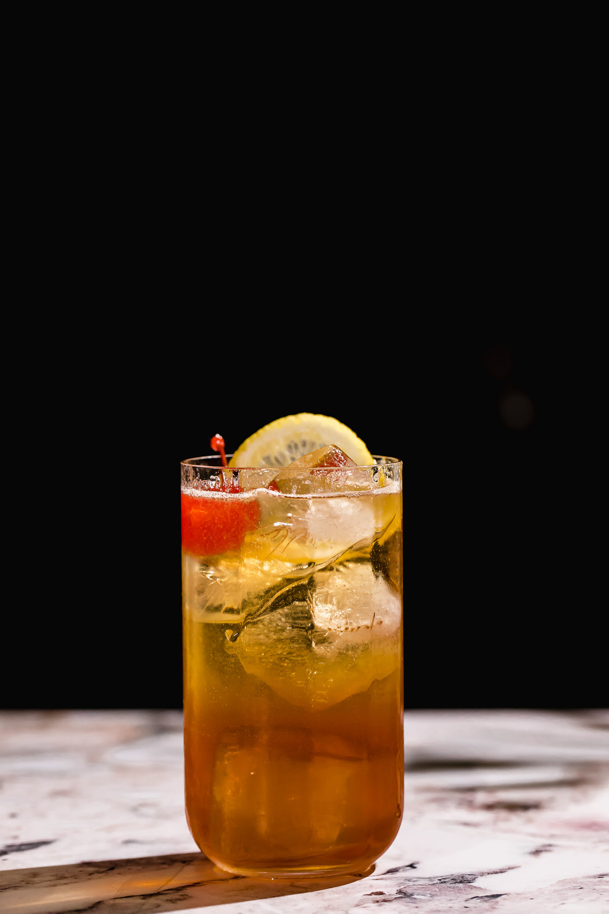 A gin sling in a glass, garnished with a lemon slice and cherry rests on a marble surface against a dark background.