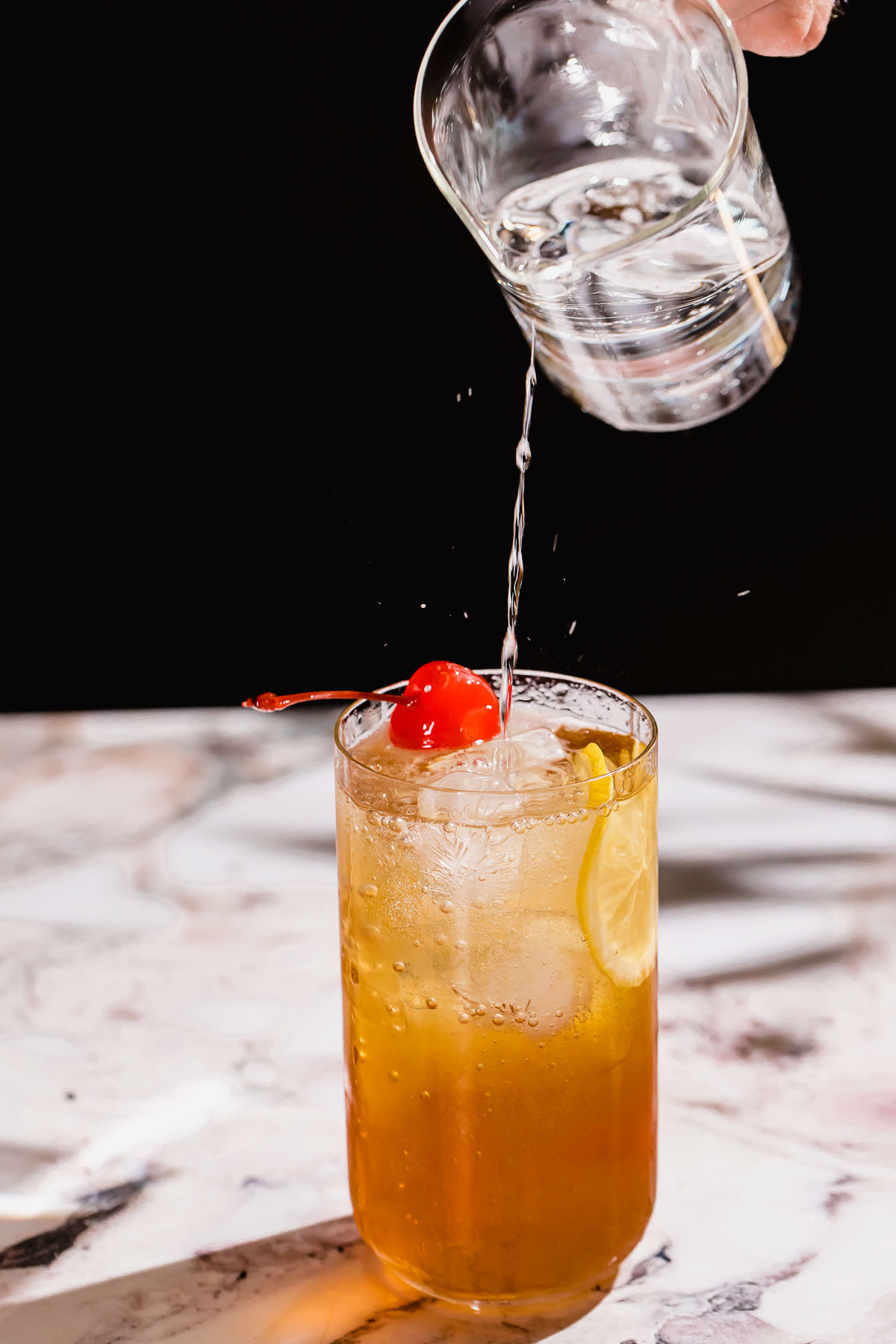 A hand pours tonic water into a cherry and lemon slice garnished iced cocktail on a marbled surface.