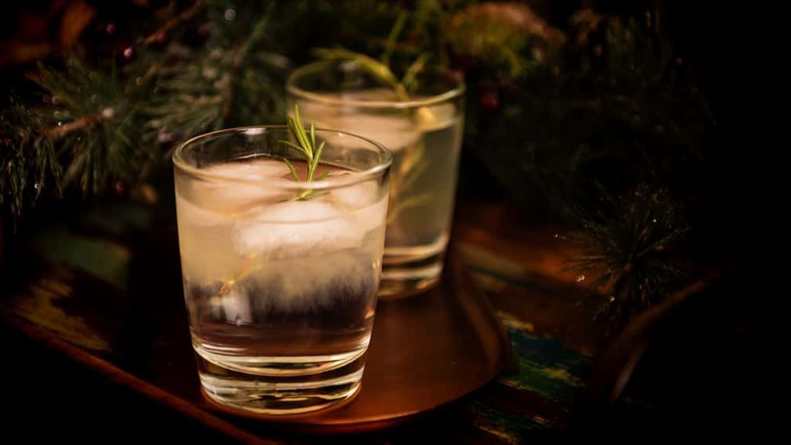 Two rosemary-garnished cocktail glasses with ice cubes rest on a dark wooden surface, surrounded by green foliage.