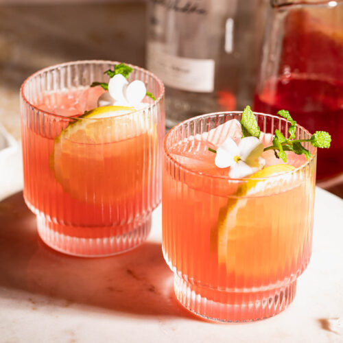 Two pink vodka lemonade cocktails with lemon and mint in ridged glasses sit on a marble surface, accompanied by a vodka bottle and liquid-filled carafe in the background.