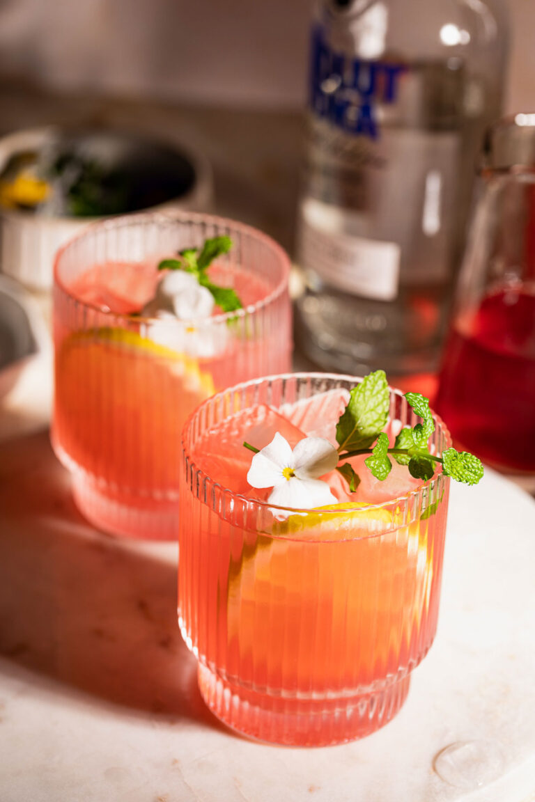 Two pink vodka lemonade cocktails in ribbed glasses garnished with lemon slices, mint, and white flowers are set before a vodka bottle and small pitcher.