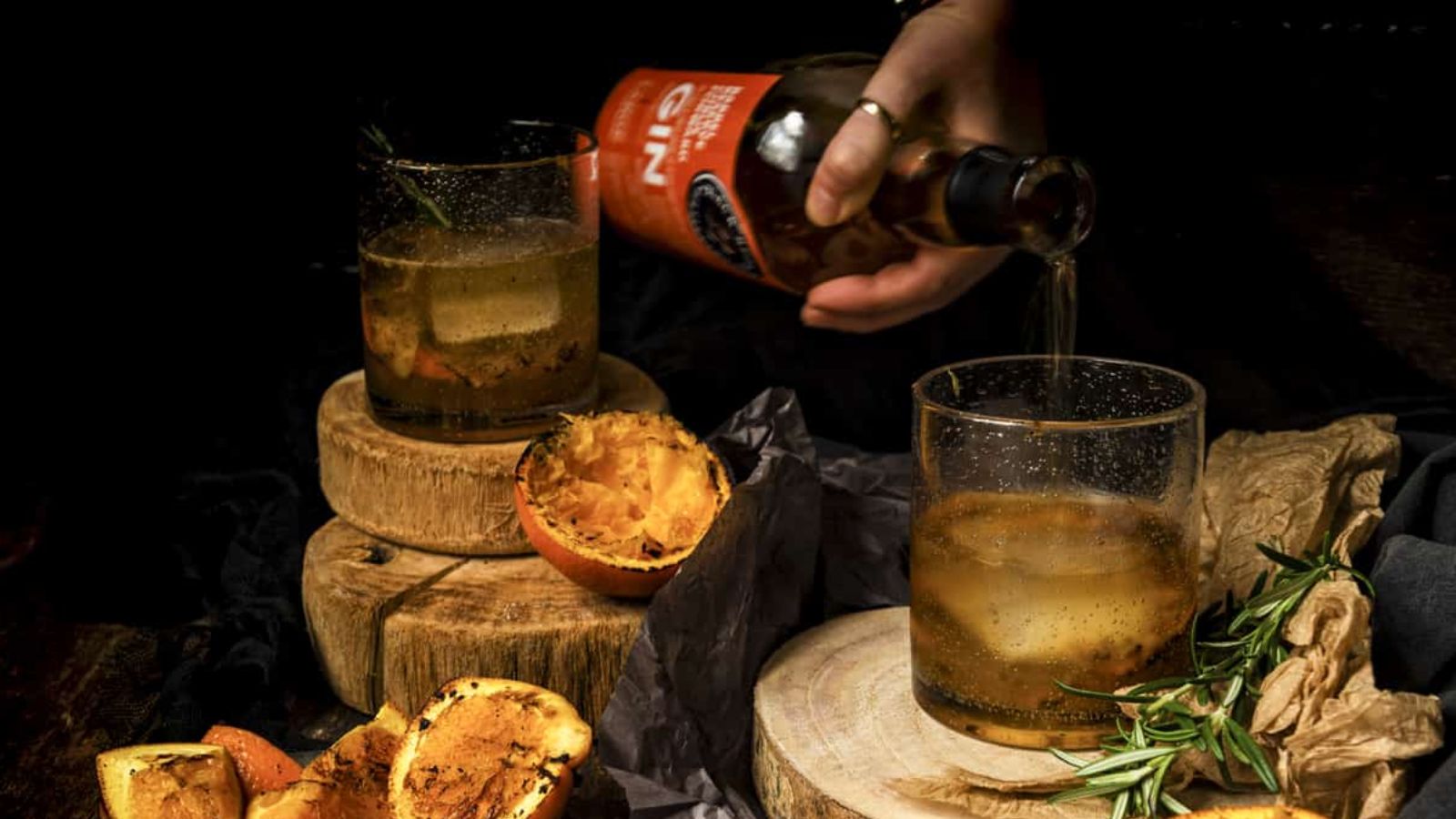 A person pours gin over ice in a glass on a wooden surface surrounded by cut oranges and rosemary.