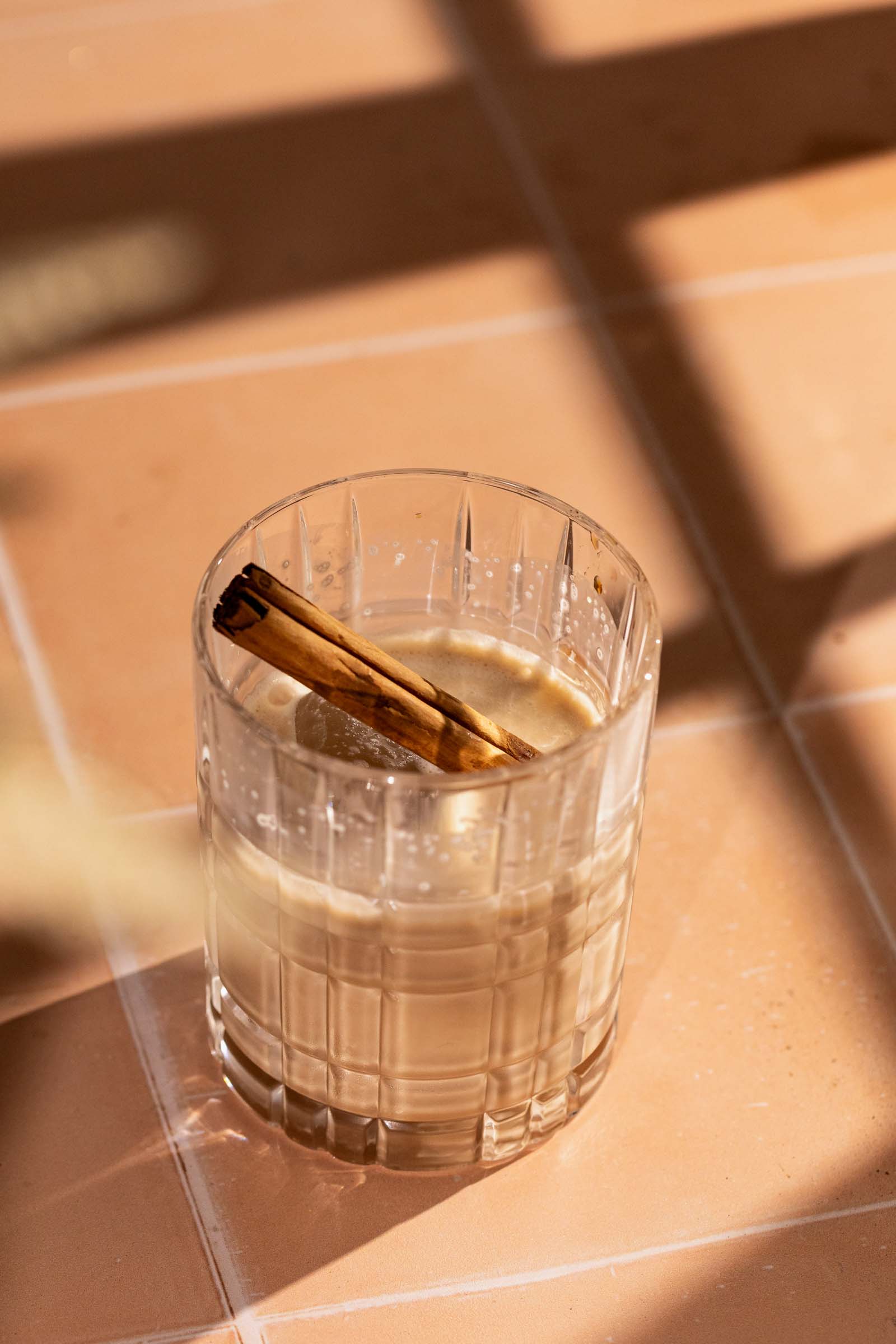 A white russian with a cinnamon stick sits on a sunlit tiled surface.