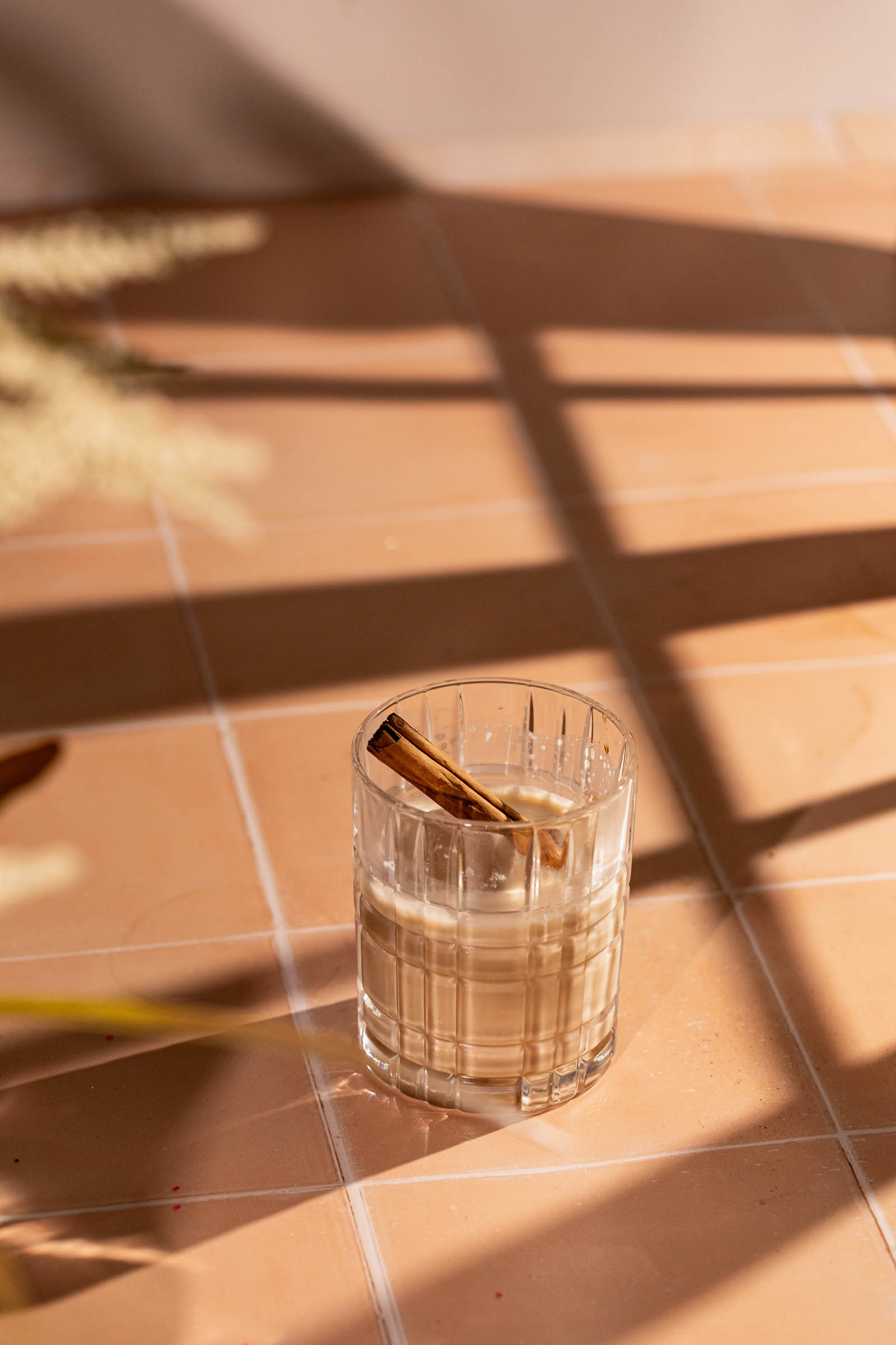 A glass of white russian cocktail garnished with a cinnamon stick sits on a tiled surface with shadows cast across it.