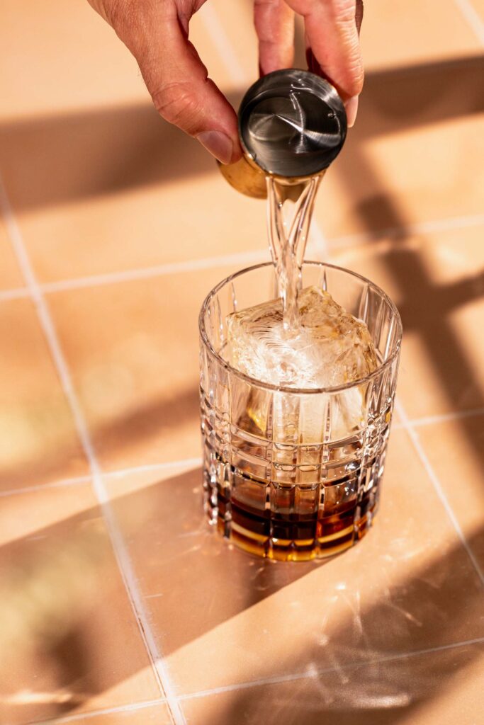 A hand pours vodka from a small cup into a glass with a large ice cube on a tiled surface.