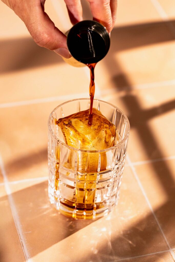 A hand pours dark coffee liqueur over a large ice cube in a glass on a tiled surface.